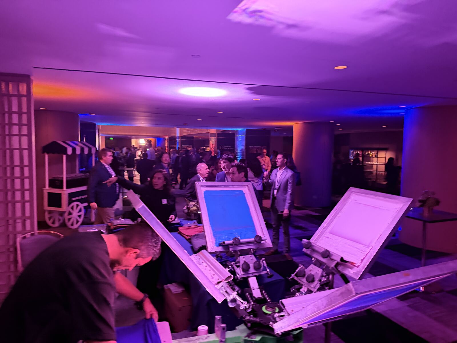 Interior event space with live printing display stations and attendees browsing decorated apparel under dramatic purple and orange lighting