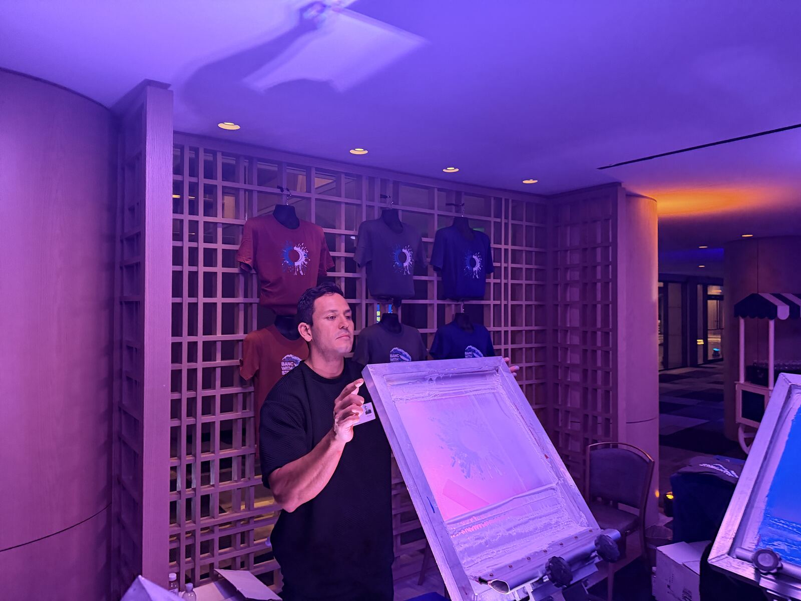 Man demonstrating DTF printing at an event booth with decorated t-shirts on a display stand under purple accent lighting