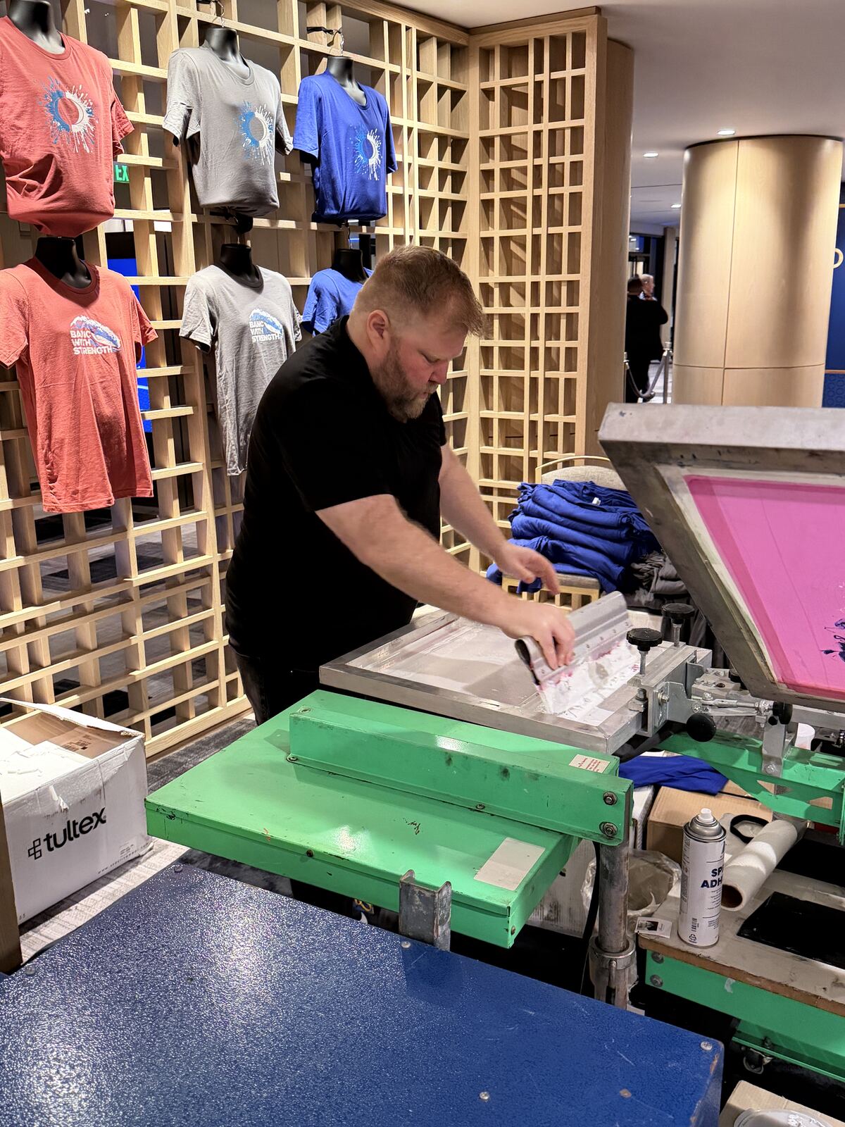Man operating a DTF heat-press machine at an indoor event booth with colorful decorated t-shirts displayed on shelving